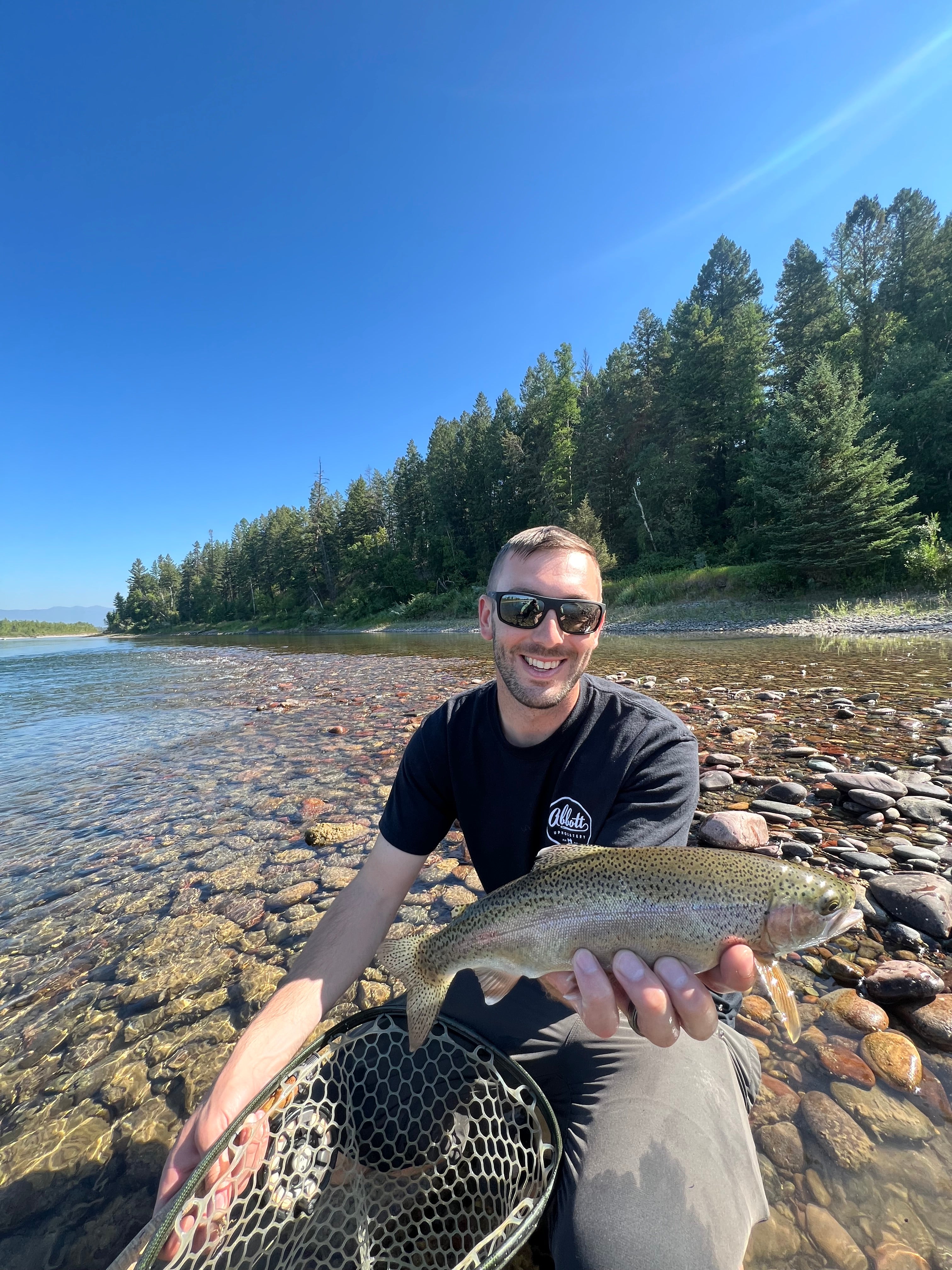 Man holding a rainbow trout on the flathead river with clear blue sky and trees in the background